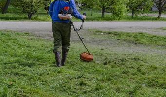 en närbild av en arbetstagare i skyddande Kläder, handskar, sudd stövlar med en gas gräsklippare på de främre gräsmatta. en man klipper gräs med maskrosor på en värma solig vår dag. foto