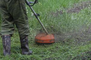 en närbild av en arbetstagare i skyddande Kläder, handskar, sudd stövlar med en gas gräsklippare på de främre gräsmatta. en man klipper gräs med maskrosor på en värma solig vår dag. foto