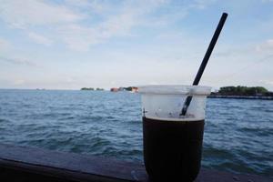 selektiv fokus på en plast kopp som innehåller svart iced kaffe med en hav se i de bakgrund foto