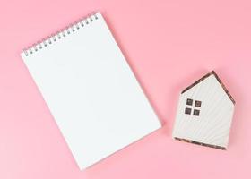 platt layout av trä- hus modell och tom sida anteckningsbok på rosa bakgrund med kopia Plats, Hem inköp planera begrepp. foto