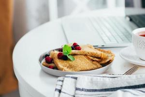 skrivbordet tabell med bärbar dator, färsk crepes pannkakor, kopp av te och honung burk. foto