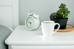 morgon- Hem rutin. vit kopp och larm klocka på bedside tabell foto