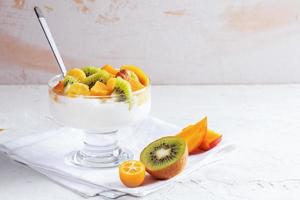glas skål på stam med yoghurt och färsk frukt är stående på vit servett på vit tabell. foto