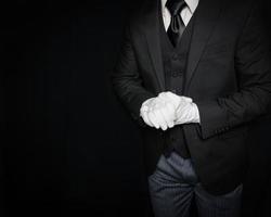 porträtt av butler eller tjänare i mörk kostym och vit handskar redo till hjälp på svart bakgrund. service industri. professionell gästfrihet och artighet foto