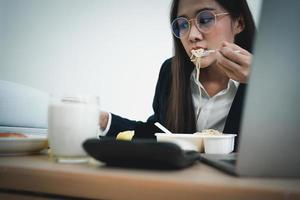 upptagen och trött affärskvinna äter spaghetti för lunch på de skrivbord kontor och arbetssätt till leverera finansiell uttalanden till en chef. överarbetad och ohälsosam för redo måltider, burnout begrepp. foto