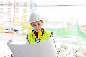 asiatisk ingenjör eller ung kvinna arkitekt satte på sig en hjälm för säkerhet och titta på ritningen för att inspektera byggarbetsplatsen på en byggarbetsplats. smart arbetande kvinna koncept. foto
