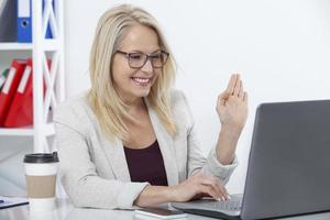 Lycklig affärskvinna med glasögon arbetssätt, chatta med bärbar dator på kontor foto