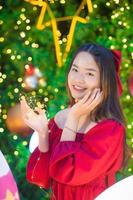 asiatisk skön kvinna i röd klänning står lyckligt i hans hand han hölls en ljus i främre av de jul träd med bokeh som bakgrund i de tema av jul och ny år fester foto