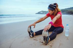 asiatiska kvinnor jogging träning på stranden. sitta ner på stranden fitness relax med stretch ben och stretch arm. foto