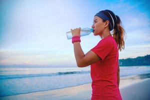 asiatiska kvinnor jogging träning på stranden på morgonen. koppla av med havspromenaden och dricksvatten från plastflaskorna foto