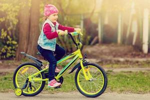 skön leende liten flicka ridning cykel i en parkera foto