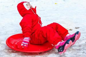 Söt leende liten flicka i henne åka skidor kostym glidning ner en små snö täckt kulle med henne släde foto