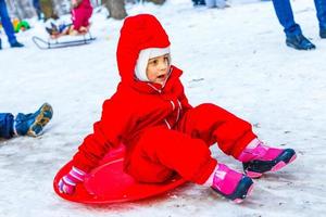 Söt leende liten flicka i henne åka skidor kostym glidning ner en små snö täckt kulle med henne släde foto