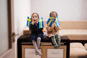 två systrar är Sammanträde på en soffa på Hem med svenska flaggor på händer. Sverige barn flickor med flagga . foto