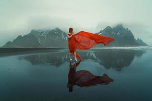 kvinna i röd klänning med vinka cape på strand naturskön fotografi foto