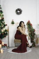 porträtt av Söt ung flicka mysigt sitta ner, leende ha på sig röd klänning i dekorerad jul levande rum inomhus foto