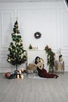 porträtt av Söt ung flicka mysigt sitta ner på de golv, leende ha på sig röd klänning i dekorerad jul levande rum inomhus foto