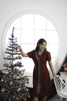 porträtt av en Söt ung flicka bär en röd klänning, leende på de kamera, stående i dekorerad jul levande rum inomhus foto