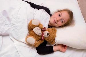 liten flicka lögner i säng med en teddy Björn och ler foto