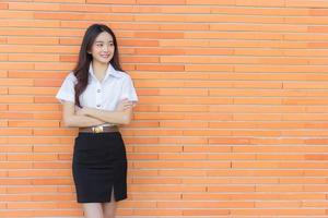 porträtt av ett vuxen thai studerande i universitet studerande enhetlig. skön asiatisk flicka stående leende lyckligt och självsäkert med henne vapen korsade på en tegel bakgrund. foto