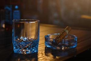 glasögon av whisky med rökning cigarr och is kuber på trä- tabell foto