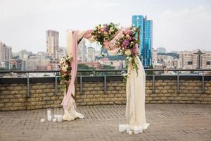 skön bröllop ceremoni utomhus. bröllop båge tillverkad av trasa och vit och rosa blommor på de tak mot de bakgrund av en stor stad . gammal dörrar, rustik stil. foto