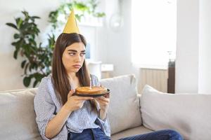 virtuell fest under nedstängning. upprörd och frustrerad enda afrikansk amerikan kvinna i fest hatt fira födelsedag uppkopplad. lady framställning video konferens, innehav kaka med ljus, Sammanträde på de golv foto