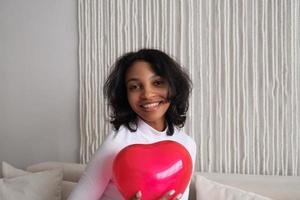 närbild porträtt av ung afrikansk amerikan kvinna innehav röd hjärta formad ballong Sammanträde på soffa på Hem. fira alla hjärtans dag, kärlek dag, kvinnors dag, födelsedag. foto