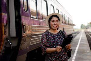 Lycklig asiatisk mitten åldrig kvinna resande är på järnväg station, bär hatt, innehar smart läsplatta och ler. begrepp, resa förbi tåg i thailand kan bok eller förbi biljett uppkopplad. foto