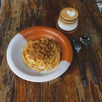 friska tallrik av italiensk spaghetti toppade med en gott tomat och jord nötkött bolognese sås och färsk basilika på en rustik brun trä- tabell. eras med cappuccino foto