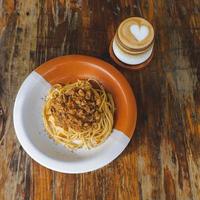 friska tallrik av italiensk spaghetti toppade med en gott tomat och jord nötkött bolognese sås och färsk basilika på en rustik brun trä- tabell. eras med cappuccino foto