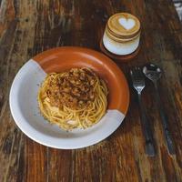 friska tallrik av italiensk spaghetti toppade med en gott tomat och jord nötkött bolognese sås och färsk basilika på en rustik brun trä- tabell. eras med cappuccino foto