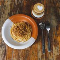 friska tallrik av italiensk spaghetti toppade med en gott tomat och jord nötkött bolognese sås och färsk basilika på en rustik brun trä- tabell. eras med cappuccino foto