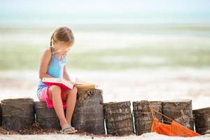 liten bedårande flicka som läser bok under den tropiska vita stranden foto