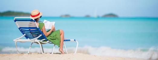 ung kvinna läsning bok på schäslong på de strand foto