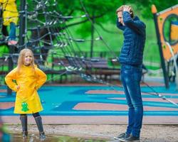 liten flicka njut av regnig väder i utomhus- lekplats, medan pappa är suprised foto