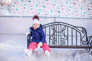 liten flicka Sammanträde på en bänk i de skridskoåkning rink foto