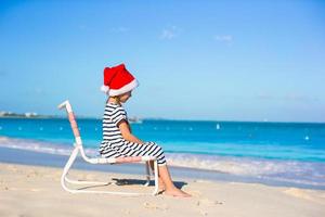 liten förtjusande flicka i röd santa hatt på strand stol foto