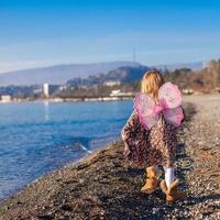 förtjusande liten flicka med fjäril vingar löpning längs de strand i en vinter- solig dag foto