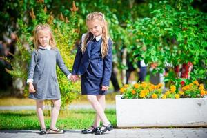 två söt smilling liten flickor Framställ i främre av deras skola. förtjusande liten barn känsla mycket upphetsad handla om gående tillbaka till skola foto