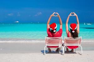 Lycklig romantisk par i röd santa hattar på strand framställning hjärtan foto
