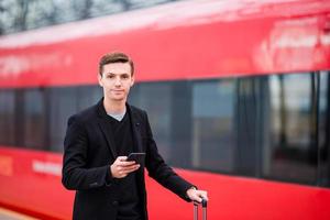 ung caucasian man med smarphone och bagage på station reser förbi tåg foto
