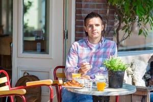 Lycklig ung man i utomhus Kafé på europeisk stad har frukost äter mysli foto
