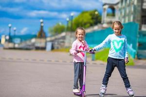 två liten flickor vält skridskoåkning och ridning en skoter i de parkera utomhus- foto