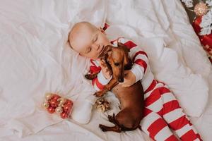 liten pojke i röd och vit pyjamas är liggande i säng på en vit ark med en hund. jul morgon. friska sömn av de barn. Plats för text. hög kvalitet Foto