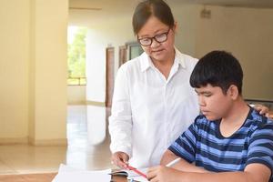 ung asiatisk pojke är håller på med hans uppdrag och lyssnande till hans kvinna äldre lärarens råd handla om skola projekt, vuxen hjälper barn till do skola projekt begrepp. foto