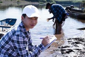 ung asiatisk pojke innehar transparent rör som har exempel vatten inuti till do de experimentera och ph nivå mått som hans skola projekt arbete med hans vänner Bakom på de flod var han levde. foto