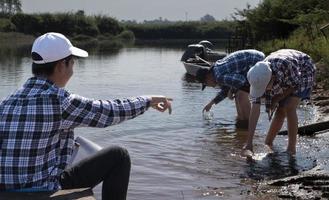ung asiatisk pojke innehar transparent rör som har exempel vatten inuti till do de experimentera och ph nivå mått som hans skola projekt arbete med hans vänner Bakom på de flod var han levde. foto
