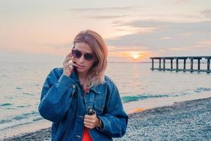 utomhus- mode porträtt av eleganta flicka bär trendig solglasögon och jeans jacka på de strand. foto