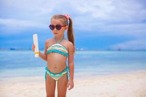 liten förtjusande flicka i baddräkt innehar solbränna lotion flaska foto